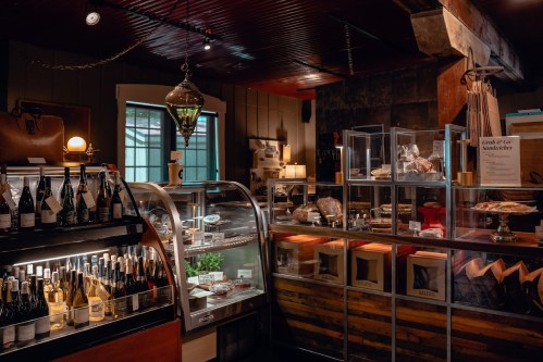 A cozy shop interior featuring a display case with pastries, shelves of wine, and rustic wooden accents.
