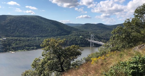 A scenic view of a river surrounded by green mountains and a bridge, under a partly cloudy sky.