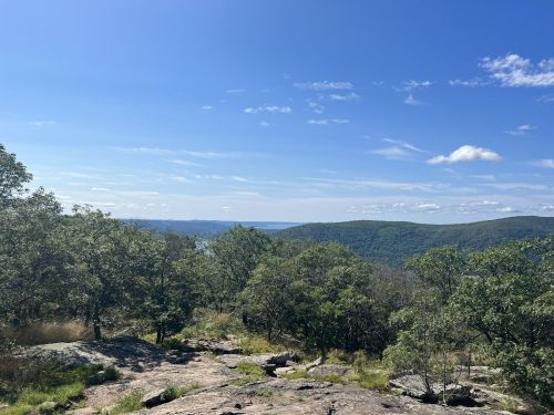 A scenic view of rolling hills and a lake under a clear blue sky, framed by trees and rocky terrain.