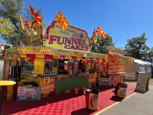 Colorful food stand with signs for funnel cakes, lemonade, and coffee, surrounded by festive flags and decorations.