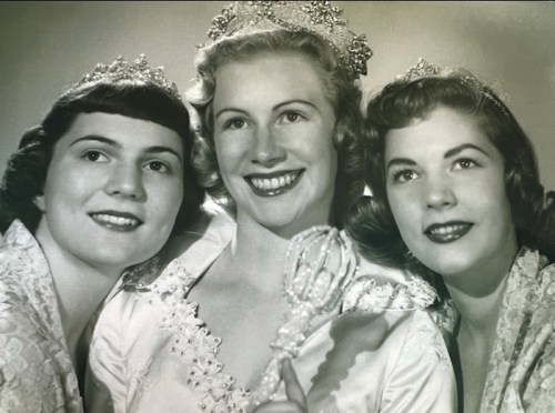 Three smiling women in tiaras and elegant dresses, posing together with a scepter, in a vintage black-and-white photo.