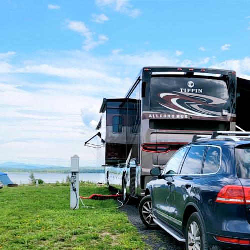 A Tiffin Allegro RV parked by a lake, with a car nearby and a scenic view of clouds and mountains in the background.