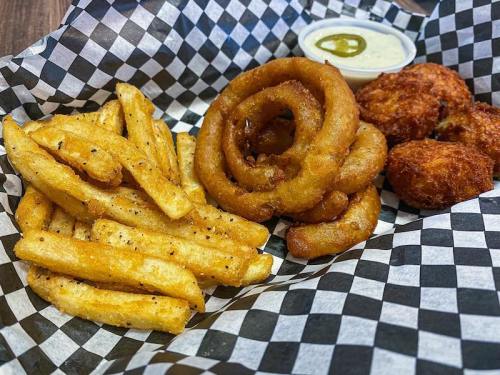 A basket of crispy fries, onion rings, and fried snacks with a side of dipping sauce and a jalapeño slice.