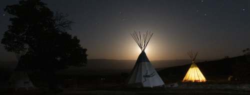 Silhouetted tipis under a starry night sky, illuminated by a glowing moonrise in the background.
