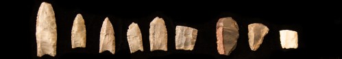 A row of various stone tools, including sharp-edged blades and points, displayed against a black background.