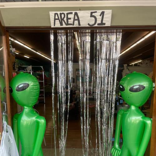 Two green alien figures stand at the entrance of a shop labeled "AREA 51," with shiny streamers hanging above.