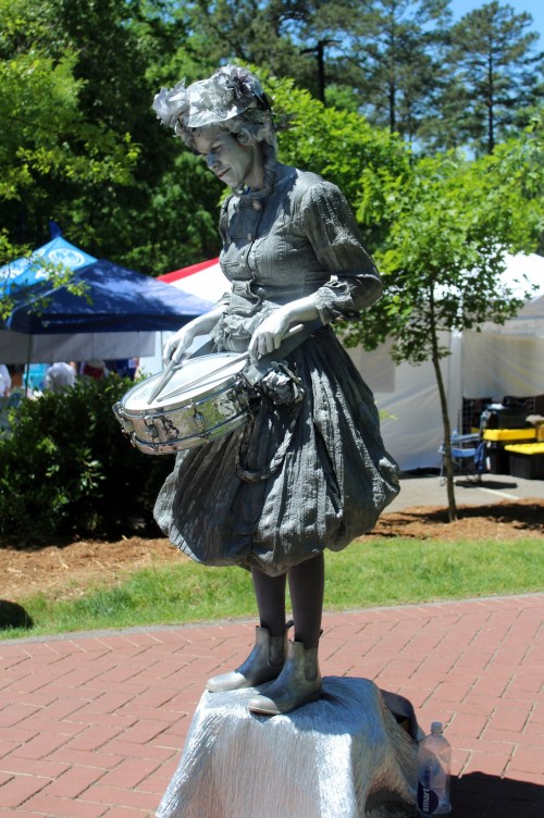A silver-painted statue of a woman in a dress, holding a drum, stands on a pedestal at an outdoor event.