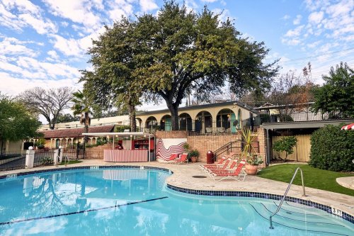 A serene pool area with lounge chairs, a tree, and a building in the background under a partly cloudy sky.