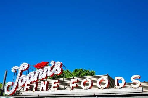 Sign for "Joan's Fine Foods" against a clear blue sky, featuring bold red and white lettering.