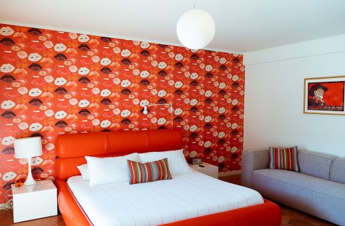 Brightly decorated bedroom featuring an orange bed, colorful patterned wallpaper, and a cozy gray sofa.