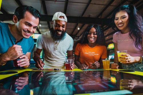 Four friends enjoy drinks and play an arcade game, smiling and having fun in a vibrant gaming venue.