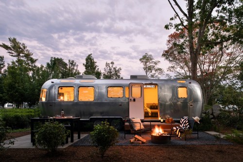 A silver Airstream trailer at dusk, surrounded by trees, with a cozy outdoor seating area and a fire pit.