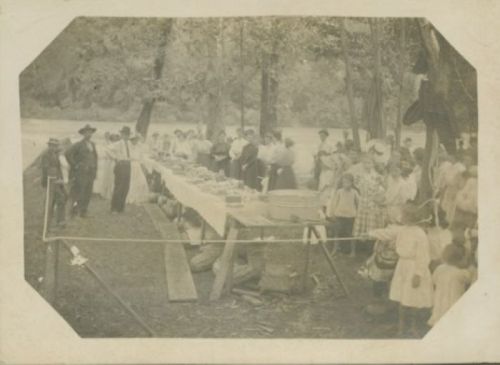 A historical outdoor gathering with a long table of food, surrounded by people in period clothing near a river.