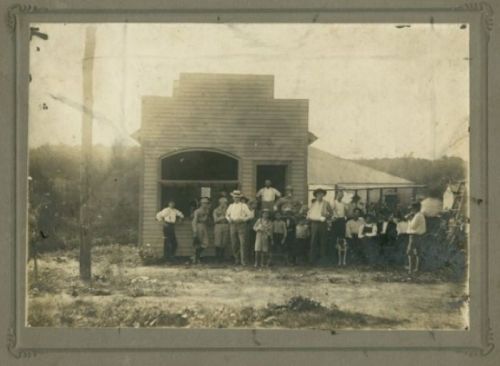 A historic black-and-white photo of a group of people standing outside a wooden building in a rural setting.