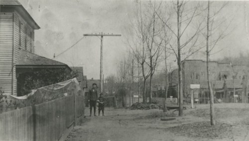 Black and white photo of a street scene with a house, a power line, and two figures standing near a fence.