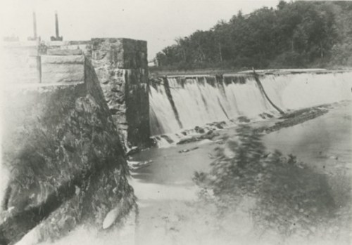 Black and white photo of a dam with water cascading over its edge, surrounded by trees and a river.