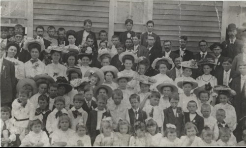 A large black-and-white group photo of men, women, and children, all dressed in early 20th-century clothing.