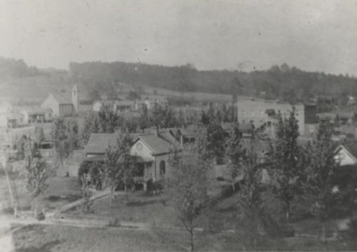 A historic black-and-white photo of a small town with houses and trees, set against a hilly landscape.