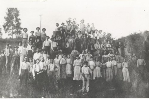 A large group of men, women, and children posed together outdoors, dressed in early 20th-century clothing.
