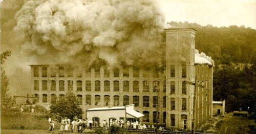 Historic photo of a factory engulfed in smoke, with people gathered nearby, witnessing the fire.