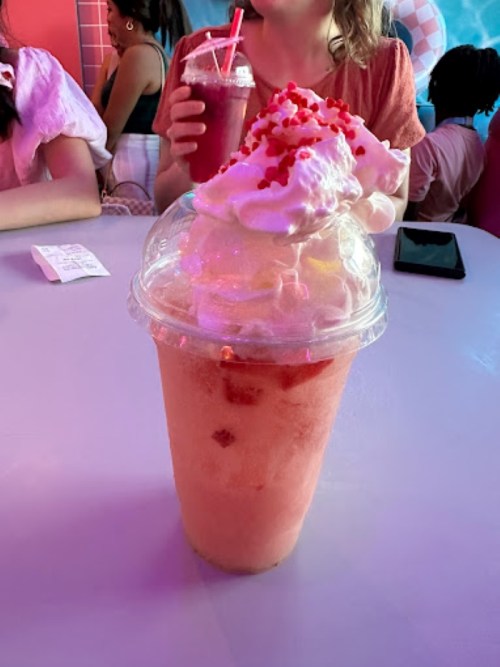 A colorful drink topped with whipped cream and sprinkles, served in a clear cup, with people in the background.