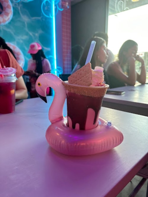 A colorful dessert served in a flamingo-shaped cup, with a wafer and whipped cream, on a table in a lively café.