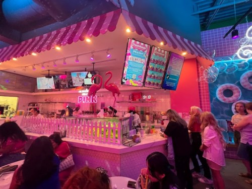 A colorful dessert shop with a pink theme, featuring flamingo decorations and a vibrant menu display. Customers are enjoying treats.