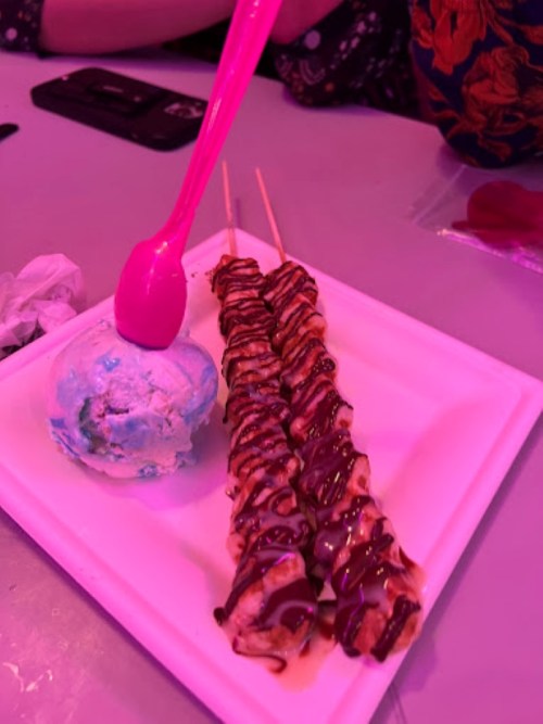 A plate with two chocolate-drizzled skewers and a scoop of blue ice cream, with a pink spoon resting on top.