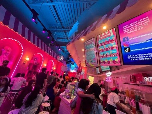 A vibrant dessert shop filled with people, featuring colorful decor, neon lights, and a menu display on the wall.