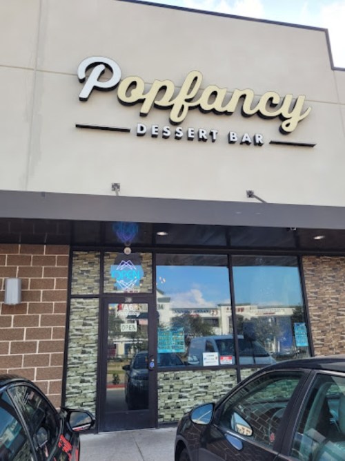 Sign for "Popfancy Dessert Bar" on a storefront, with cars parked outside and a clear blue sky above.