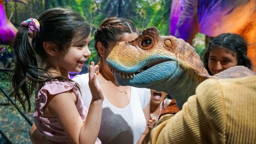 A young girl smiles and waves at a lifelike dinosaur puppet, surrounded by excited onlookers in a vibrant setting.