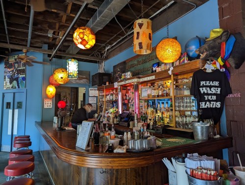 A vibrant bar with colorful pendant lights, a wooden counter, and a sign that says "Please wash your hands."
