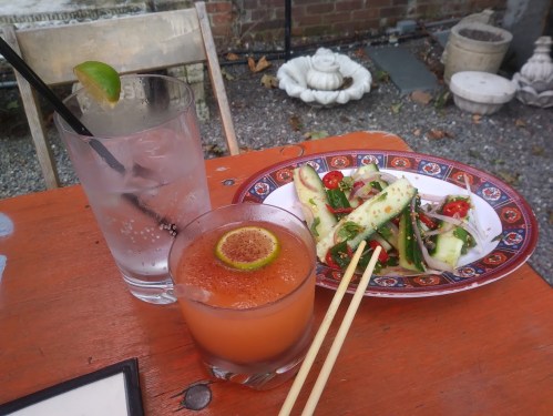 A table with a cocktail, a glass of water, and a plate of fresh salad with vegetables, set in a garden.
