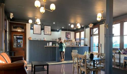 A cozy café interior with a counter, seating area, and large windows, featuring a person standing at the counter.