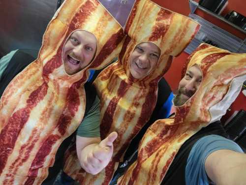 Three people wearing bacon-themed costumes pose for a selfie, smiling and giving a thumbs up.