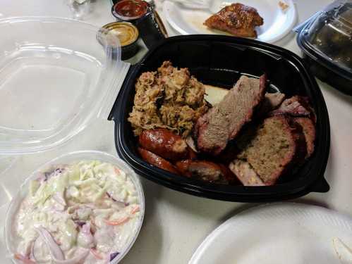 A takeout container with pulled pork, sliced brisket, sausage, and a side of coleslaw on a table.