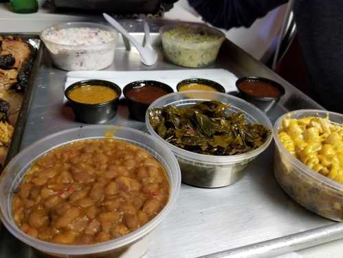 A metal tray with various dishes: baked beans, collard greens, macaroni, and three sauces in small containers.