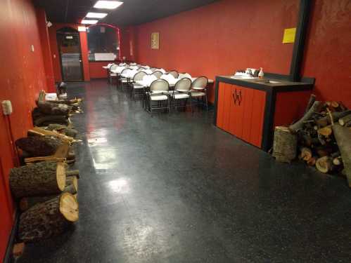 Interior of a red-walled room with a long table and chairs, and stacks of firewood along the walls.