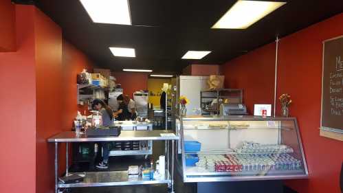 Interior of a small restaurant with a kitchen area, display case, and staff preparing food. Bright red walls and black ceiling.