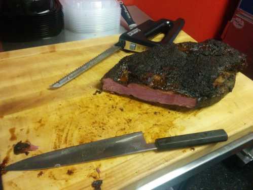 A smoked brisket on a wooden cutting board, with a knife and saw nearby, showcasing a dark, charred exterior.