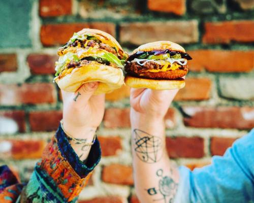 Two hands holding delicious burgers against a brick wall, showcasing vibrant toppings and sauces.
