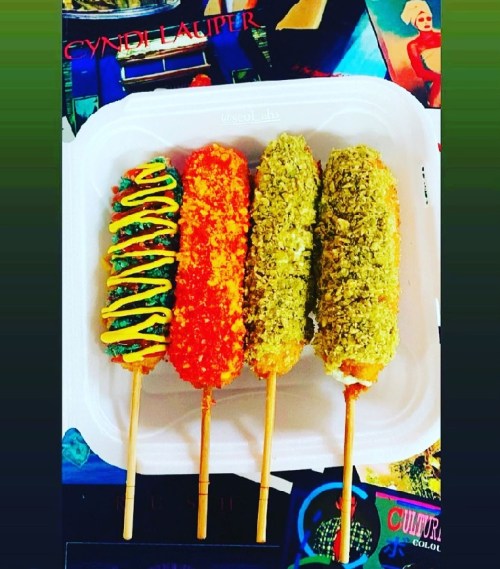Four colorful corn dogs on sticks, topped with mustard, ketchup, and green herbs, served on a white plate.