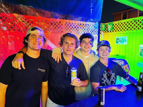 Four young men pose together at a colorful outdoor bar, smiling and enjoying drinks under vibrant lighting.
