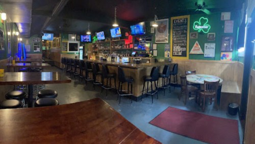 A spacious bar with a wooden counter, high stools, multiple TVs, and tables, featuring green decor and a cozy atmosphere.