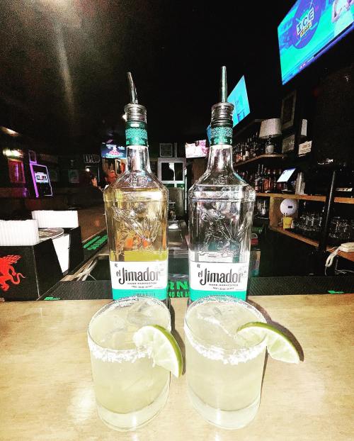 Two margaritas with lime wedges sit on a bar, with two bottles of El Jimador tequila in the background.