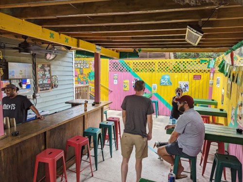 A colorful outdoor bar scene with patrons sitting at tables and a bartender behind the counter. Brightly painted walls.