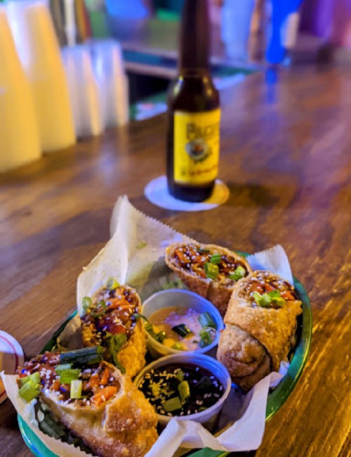 A plate of crispy spring rolls with dipping sauces, set on a wooden bar with a beer bottle in the background.