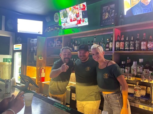 Three smiling bartenders pose together behind a bar, with drinks and screens in the background.