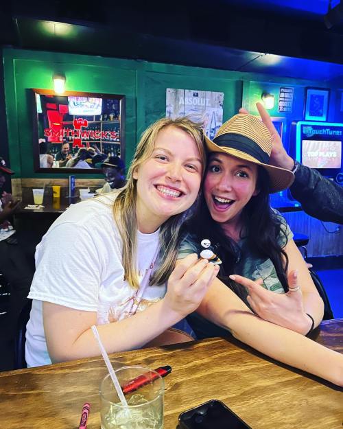 Two smiling women pose together at a bar, one holding a small figure, with colorful lights in the background.