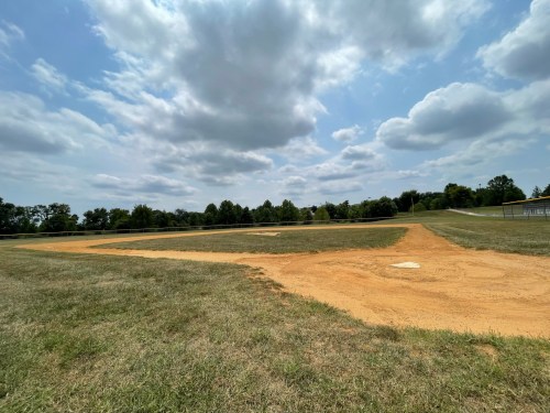A baseball field with a dirt infield, grassy outfield, and a cloudy sky overhead.